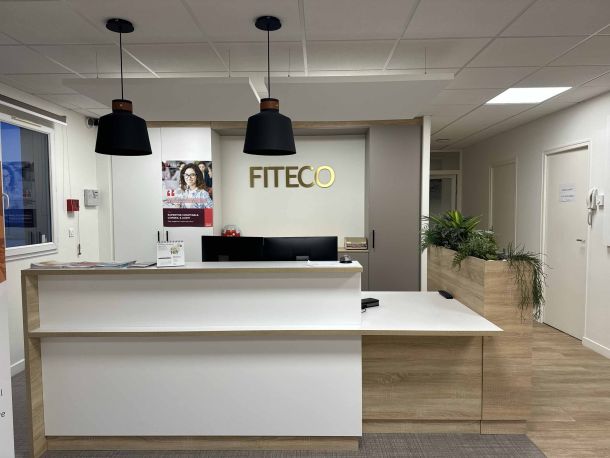Borne d’accueil moderne avec un comptoir en bois clair et blanc, bac à plantes intégré, suspensions noires au plafond, et logo FITECO en lettres dorées sur le mur, dans un bureau professionnel lumineux.